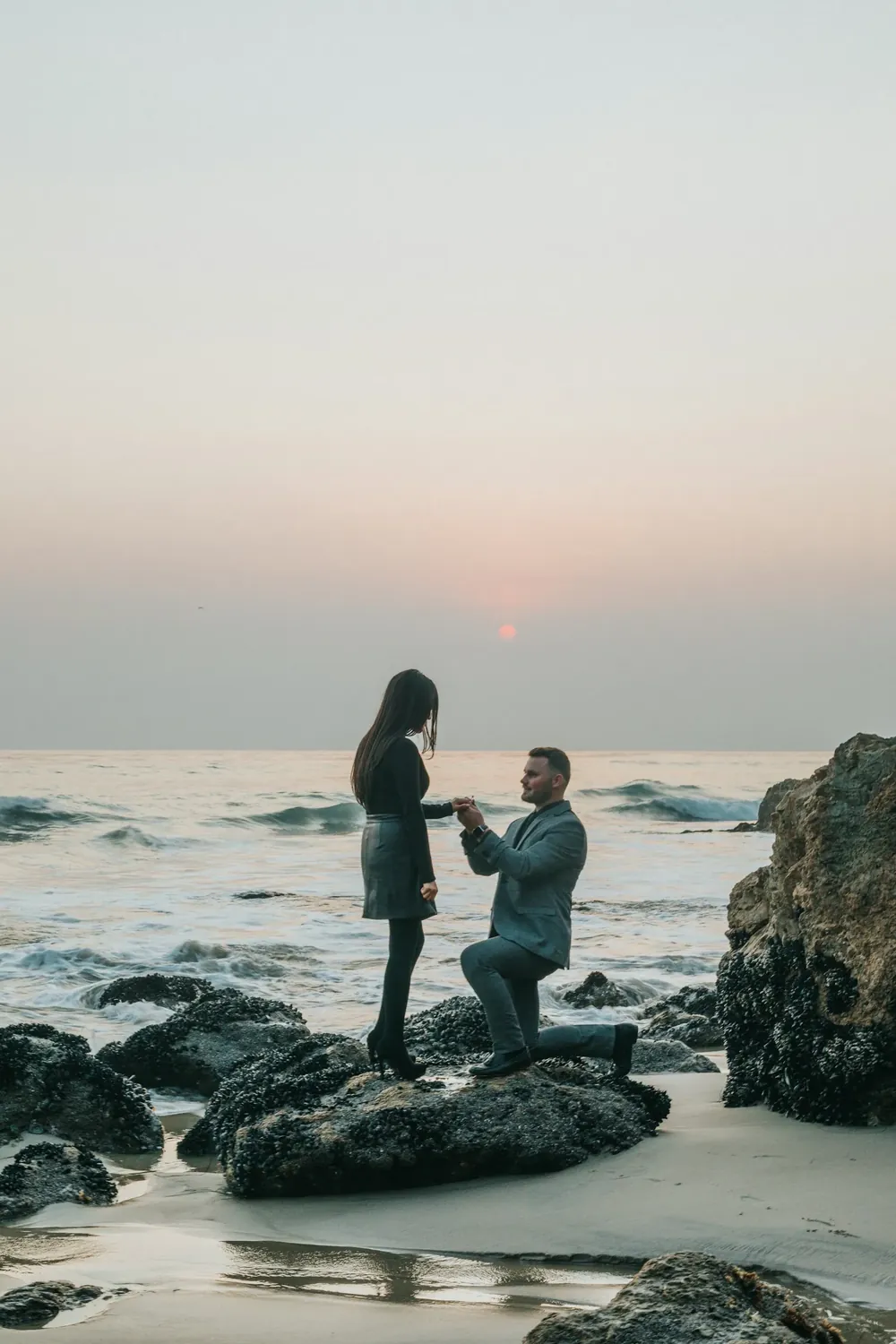 A man on one knee proposing on a rocky shore at dusk